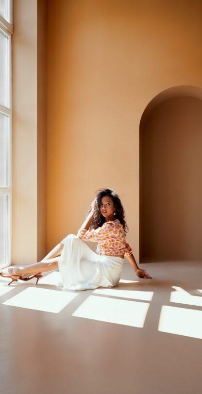Charming young lady with dark hair in flower printed blouse, white long skirt and high heels sitting on the floor near big panoramic window with sunlight. Sexy model posing on camera in studio.