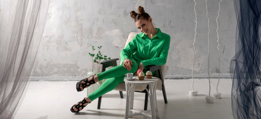 Cute girl smiles and drinks coffee with cupcakes in studio. Young woman in green suit poses on gray background.